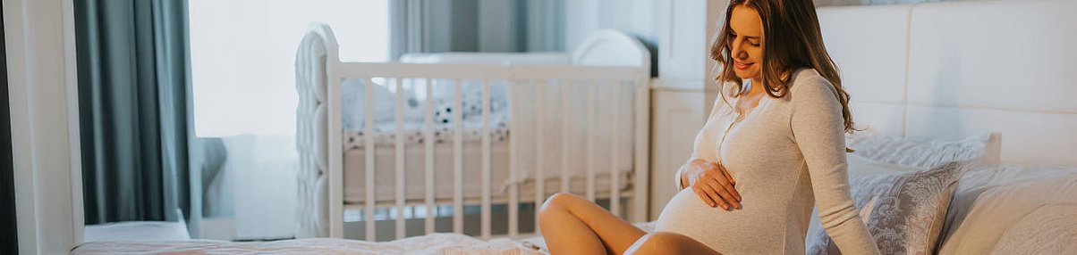 Schwangere Person sitzt auf einem Bett in einem Schlafzimmer mit Babybett im Hintergrund