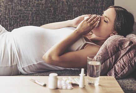 Eine schwangere Frau liegt auf einem Sofa und hält sich ein Taschentuch an die Nase. Vor ihr auf einem Tisch stehen ein Glas Wasser, verschiedene Medikamente, darunter Tabletten und Nasenspray.