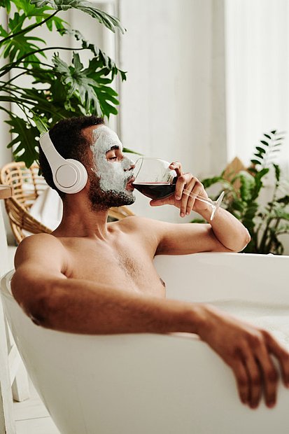 Eine Person sitzt entspannt in einer Badewanne, trägt weiße Kopfhörer und eine Gesichtsmaske. In der Hand hält sie ein Glas Rotwein. Im Hintergrund sind grüne Pflanzen und Möbel aus Naturmaterialien zu sehen – die Szene vermittelt Ruhe, Wellness und Erholung im eigenen Zuhause.