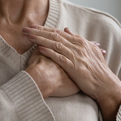 Nahaufnahme von zwei Händen, die über der Brust verschränkt liegen. Die Person trägt einen hellen, gerippten Pullover. Der Bildausschnitt konzentriert sich auf Hände und Oberkörper und symbolisiert möglicherweise Selbstfürsorge oder Beschwerden im Brustbereich – passend zum Thema Herz und Erkältung.