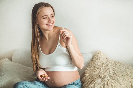 Eine schwangere Frau sitzt auf einem Sofa, trägt ein weißes Top und blaue Jeans. In der einen Hand hält sie Nüsse, mit der anderen isst sie eine. Neben ihr liegen Kissen, darunter ein flauschiges beiges Kissen.