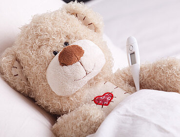 Ein Teddybär hat Fieber und liegt mit einem Thermometer zugedeckt krank im Bett