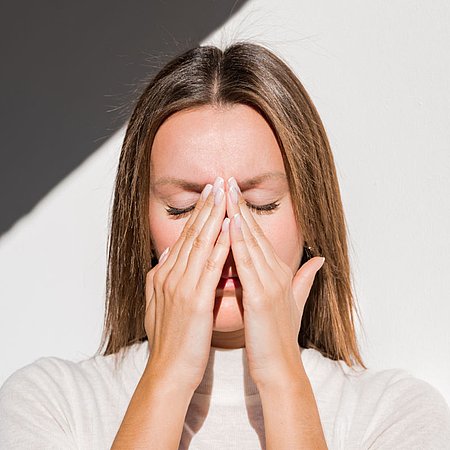 Person steht vor einer hellen Wand mit einem diagonalen Schatten und hält beide Hände vor den unteren Teil des Kopfes; gerade, schulterlange Haare und ein schlichtes, helles Oberteil sind sichtbar.