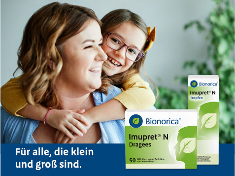 Person hält ein Kind im Arm, beide Gesichter sind unscharf dargestellt. Im Vordergrund stehen zwei Packungen des pflanzlichen Erkältungsmittels Imupret® N von Bionorica: Dragees und Tropfen. Text im Bild: „Für alle, die klein und groß sind.“