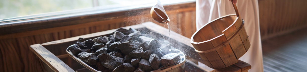 Eine Person steht in einer Sauna oder einem Saunaraum und gießt mit einer Kelle Wasser aus einem Holzeimer auf erhitzte Steine, wodurch Dampf aufsteigt.